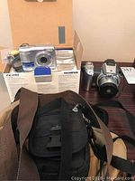 Two cameras shown with original Olympus FE-170 box including installation disks and cables, and two black camera bags with straps.