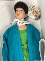 Doll in box wearing teal coat, green top, blue pants, gold heels, leopard headwear visible