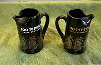 Front view of both black ceramic 100 Pipers pitchers showing graphics and text