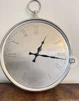 Front view of silver pocket watch style wall clock showing Roman numeral dial and black hands