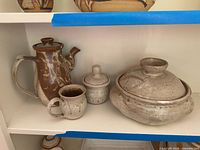 Group view of four signed stoneware pieces on shelf: coffee pot, mug, sugar jar, casserole