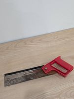 Overall view of back saw on wooden surface, showing red handle, spine and blade