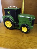 Closed tractor-shaped toolbox showing John Deere logo, wheels, handle, tape measure for scale