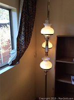 Full view of tension pole lamp in corner, revealing two illuminated milk glass shades, brass-tone pole, and wooden accents.