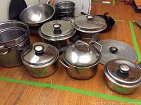 Photo showing a collection of kitchen cookware including stainless steel pots with lids, steaming baskets, mixing bowls, and cast iron skillet.