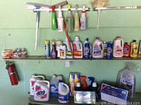 Photo of multiple shelves with a variety of household cleaning products including liquid cleaners, sprays, bleach bottles, and detergents. Two fire extinguishers mounted to the wall on the left. Boxes of plastic cutlery and Kirkland plastic cups also present.