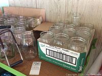 Boxes and loose Ball mason jars with metal jar lifter tool displayed on a shelf, along with blue speckled enamel canning pot visible at edge.