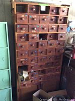 Full view of tall brown wooden cabinet with multiple small drawers, some open containing various items and blue dot stickers on drawer fronts.