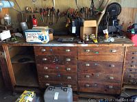 Full view of the wooden work bench with various drawers, metal handles, and a vise clamp on one side. The bench surface has wear and used marks with a few scattered items on top.