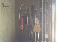 View of emergency supplies and tools hung on pegboard, including fire extinguisher, brooms, jumper cables, levels, and square ruler.