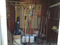 Wide view of pegboard on wall holding various garden tools including rakes, shovels, axes, saw, tamper, pruner, and pitchfork.