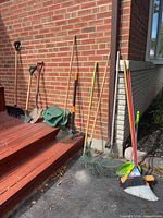 Group of garden tools leaning against brick wall including shovels, rakes, broom, dustpan