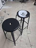 Two black metal bar stools, one seat partly grey, showing overall design