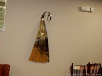 Metal wall sculpture on beige wall showing the overall shape and reflective surface with curled tip and fan-like bottom.