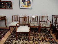 Five wooden chairs lined up against a wall on a patterned rug, two armchairs with armrests and three side chairs. Four chairs have patterned fabric cushions; one armchair has a plain light fabric cushion.