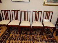 Five wooden dining chairs with upholstered seats arranged side by side showing their fronts and backs in a room with patterned carpet.