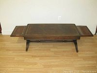 Full front view of the leather top coffee table with both extension panels visible in the open position, showing the table's overall structure including the carved pedestal legs and leather surfaces.