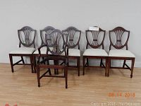 Six mahogany dining chairs with shield backs and light upholstery. One armchair with arms and five side chairs without arms all arranged against a light grey wall on a wooden floor.