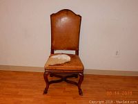 Photo showing front view of the wooden carved chair against a plain wall, highlighting the leather backrest and worn fabric on the seat.