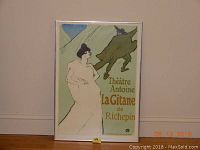 Framed art poster titled "Gitane" featuring an illustration of a woman in white and a man in green with a cat on his shoulder, displayed against a wall.