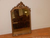 Vintage rectangular mirror with ornate gold frame and detailed carved crest at the top, placed leaning against a white wall on wooden floor.