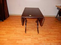 Photo of a dark wood drop leaf table with both leaves folded down, sitting on a wood floor against a white wall.