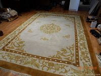 Full view of the large hand woven rug laid on a wooden floor, showing the beige and gold floral pattern with central medallion.