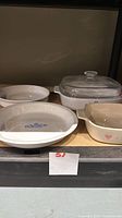 Four Corning Ware dishes on shelf: two round pie bakers, square casserole with lid, beige Forever Yours dish