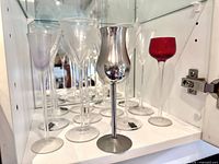 Group of cordial glasses in cabinet including clear, lilac, red, and stainless goblet