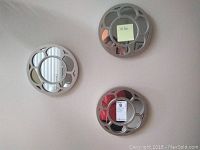 Photo of three silver colored decorative round mirrors laid flat against white background, showing flower petal-like designs in frames and 10 inch size.