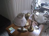 Three desk lamps on wooden table near vertical blinds and tile floor. Lamps vary in size and style, base materials mainly brass, shades include frosted and clear glass.