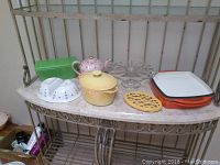 Overview of assorted serving dishes including cast iron casserole, orange grill pan, pink floral teapot, green container, white divided serving dish, clear cake stand, yellow trivet.