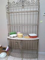 Full view of the metal baker's rack with four shelves, showing the distressed beige finish and decorative scrollwork. Includes items placed temporarily on the middle shelf but not included.