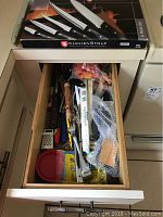 Drawer containing assorted kitchen knives and cooking utensils including peelers, graters, and packages wrapped in bubble wrap.