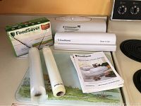 Photo shows FoodSaver Store 'n Cut bag sealer, two rolls of vacuum sealing bags, and printed instruction manuals arranged on kitchen counter.