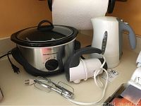 Photo showing all three appliances: Hamilton Beach slow cooker, Sunbeam hand mixer front and center, and Braun electric kettle in background.