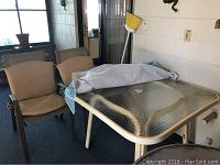 Photo showing outdoor rectangular table with glass top and three metal mesh chairs with cushions on a patio.