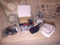 Pentax Optio A10 digital camera with box and accessories spread on couch upholstery with patterned cushions in background.