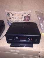 Front view showing the Canon TS6020 printer on a beige surface with a decorative pillow in the background.