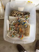 Plastic tote container holding multiple Smurf coffee mugs and water glasses with some newspaper packing.