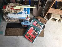 Three bags of concrete mix, a wooden handled car duster with white string mop head, a blue gallon antifreeze container, two new 6x8 tarps in sealed packaging, brown mats under.