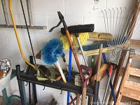 Overall view of garden tools and brushes on black plastic rack including rakes, broom, and saw