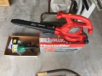 Red and black Homelite electric leaf blower vacuum resting on its original box with a green cordless shrub trimmer in a cardboard box beside it.