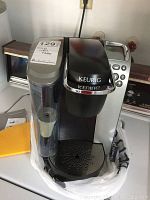 Front and top view of black and silver Keurig K-Cup coffee maker K79 model. Shows removable water reservoir on left and control buttons on right side.