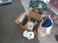 Box containing multiple picture frames, white dog statue, wooden welcome sign, and assorted bags nearby.
