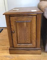 Side view of one wooden side table showing hinged door, panel design, wood grain, and knob on hardwood floor next to couch.