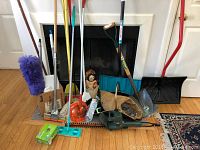 Photo showing an assortment of garden and household tools arranged on a hardwood floor in front of a fireplace, including hedge trimmer, shovels, broom, mops, and dusters.