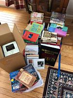 Wide view of multiple stacks and piles of paperback books on a hardwood floor, some in cardboard boxes, including a large hardcover book titled "The World's Greatest Treasury of Heart Secrets".