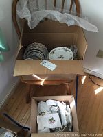 Wide image showing two open cardboard boxes containing stacked plates, bowls, and wrapped serving pieces, on wooden chair and floor