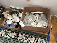 Two boxes filled with assorted cups, saucers, plates, and glass dishes placed on the floor.